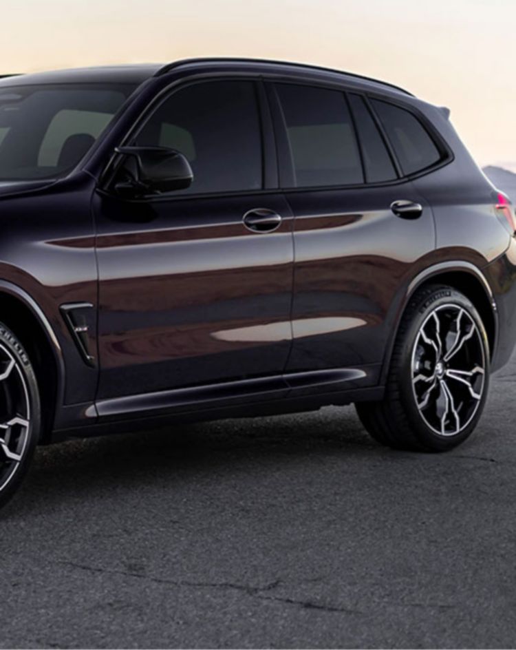 A dark-colored performance SUV parked on a cracked desert road at sunset, with mountains silhouetted on the horizon. 
