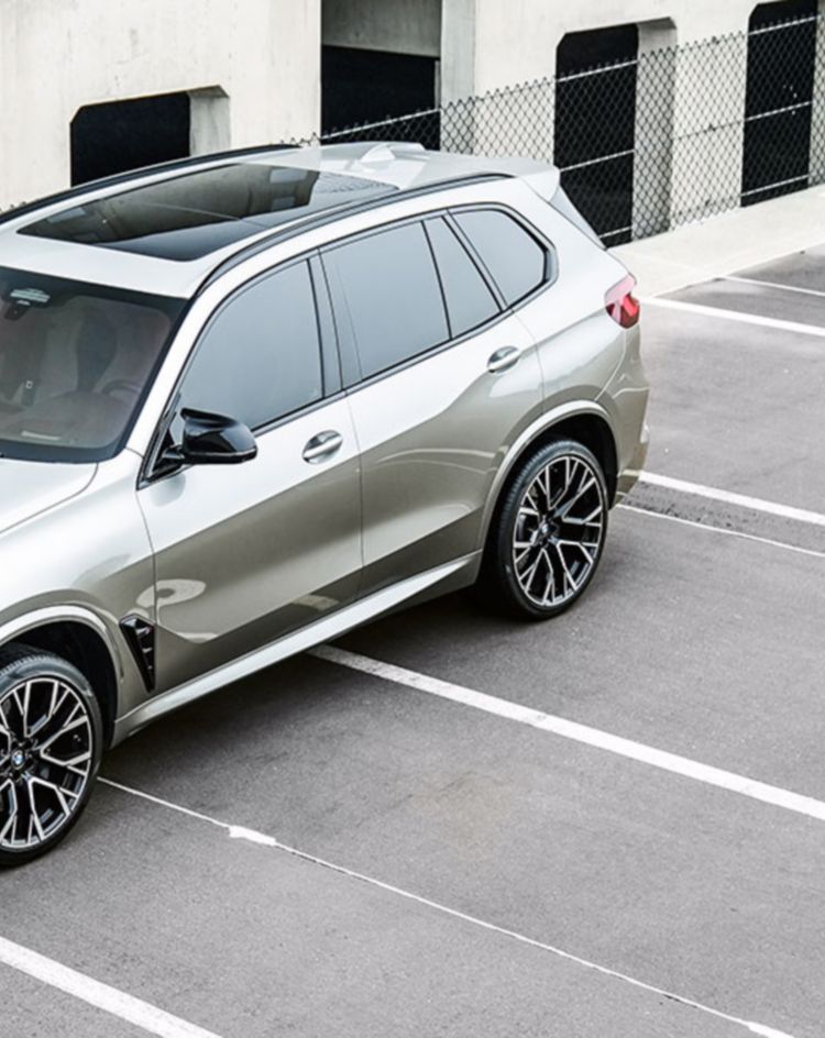 A silver performance SUV parked in an empty parking lot beside a concrete building with open garage bays and chain-link fencing. 