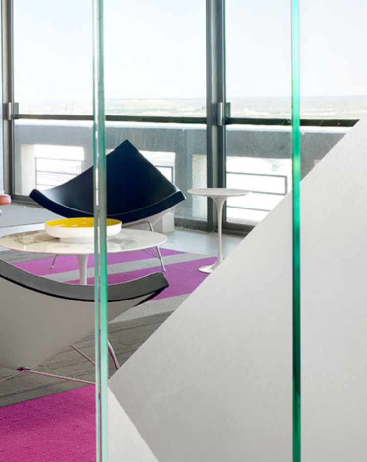 A contemporary interior space with minimalist furniture, including black lounge chairs, a colorful rug, and large floor‑to‑ceiling windows overlooking an outdoor view. 
