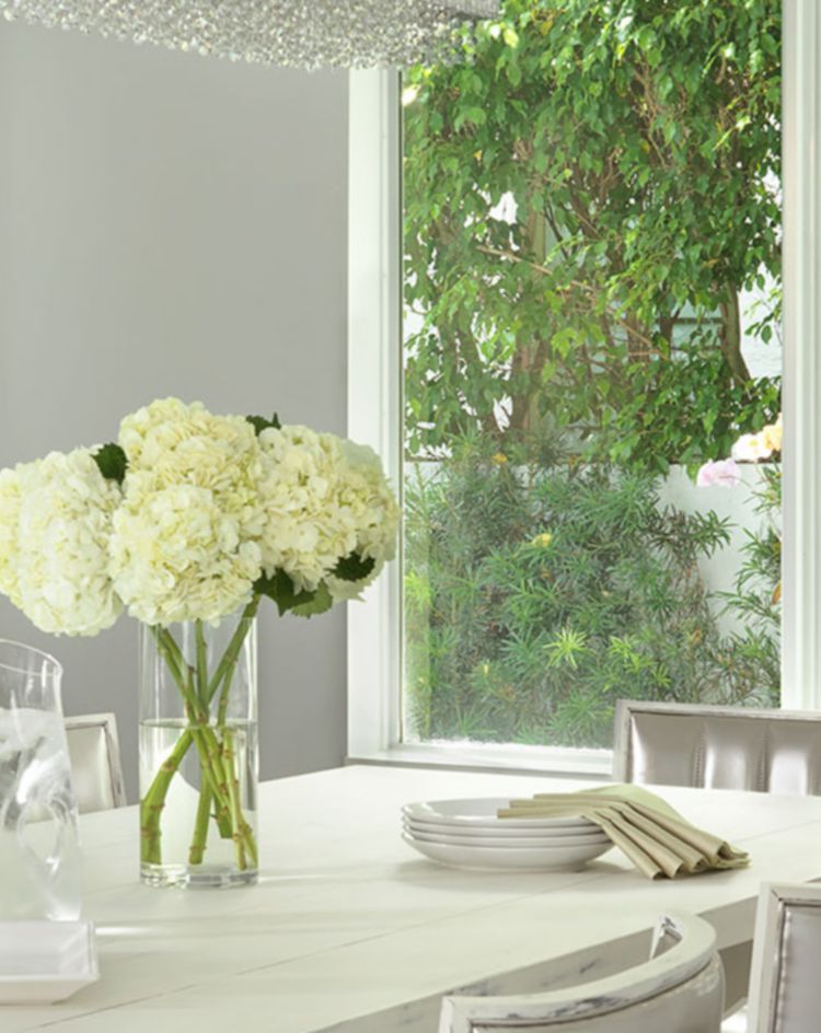 Bright dining room with a white table, neutral chairs, and large windows overlooking lush greenery. 