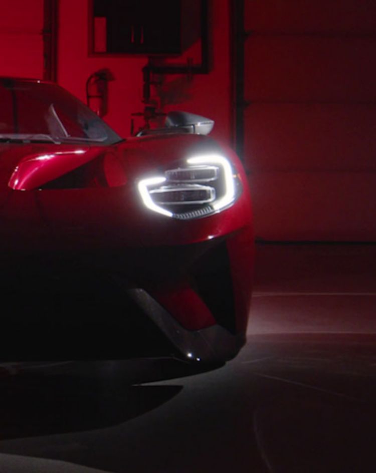 A low-lit view of a red sports car facing forward in a dark garage, with its bright LED headlights glowing against the red background. 
