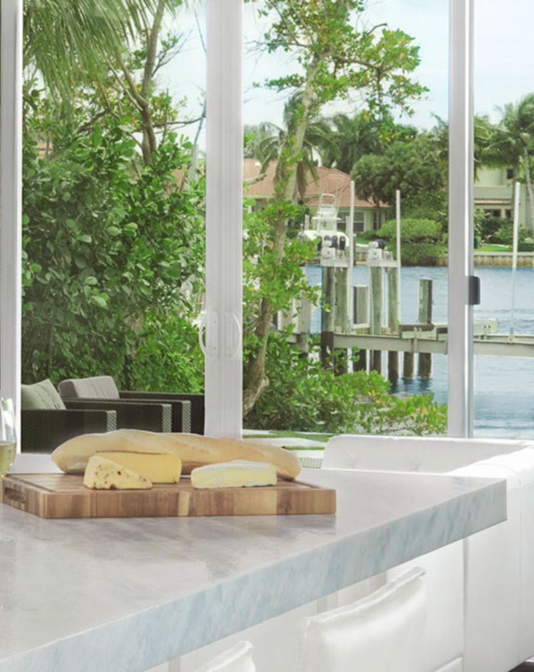 A bright modern kitchen with a large marble island, chrome pendant lights, white seating, and floor‑to‑ceiling windows overlooking lush greenery and a waterfront dock. 