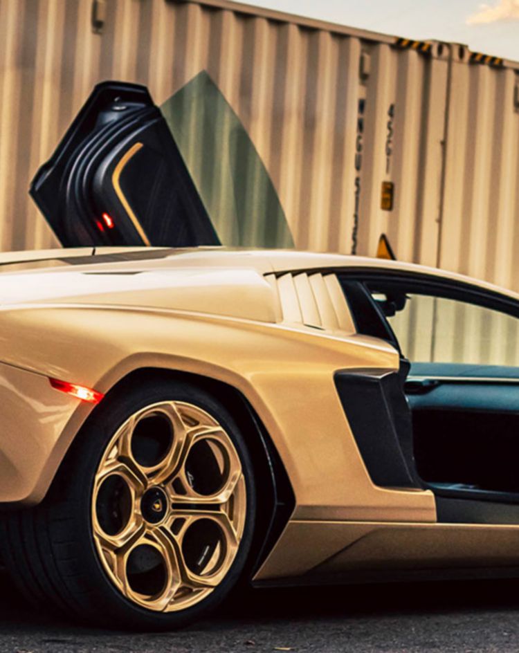 A gold-colored supercar parked on pavement with both scissor doors open, shown from a rear three‑quarter angle, with tall shipping containers in the background. 