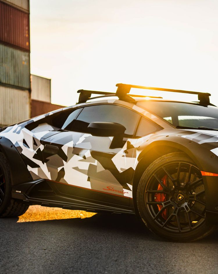 Camouflage-wrapped sports car parked near shipping containers at sunset. 