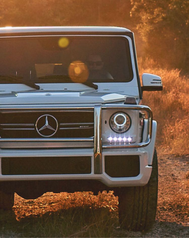 A white luxury off‑road SUV driving on a dirt trail at sunset, highlighting its rugged design, LED headlights, and premium Mercedes‑Benz styling. 