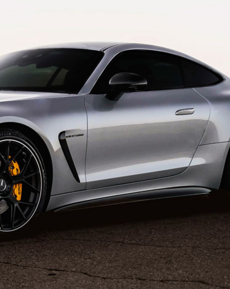 A silver sports coupe parked on a cracked desert road at sunset, with distant mountains silhouetted against the horizon. 