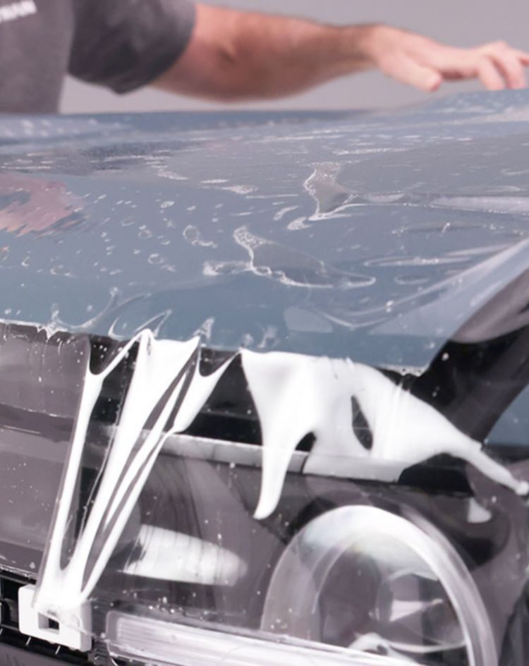 Two people applying protective film to the hood of a Bronco vehicle in a workshop. 