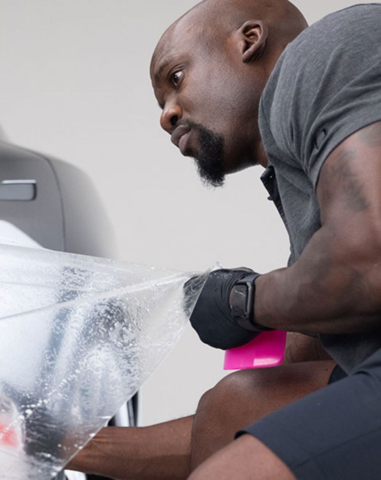 Person applying protective film to the side of a dark vehicle in a workshop. 