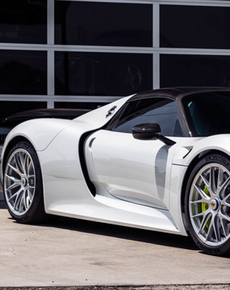 A white high‑performance sports car parked in front of a modern glass‑panel garage, shown from a front three‑quarter angle. 