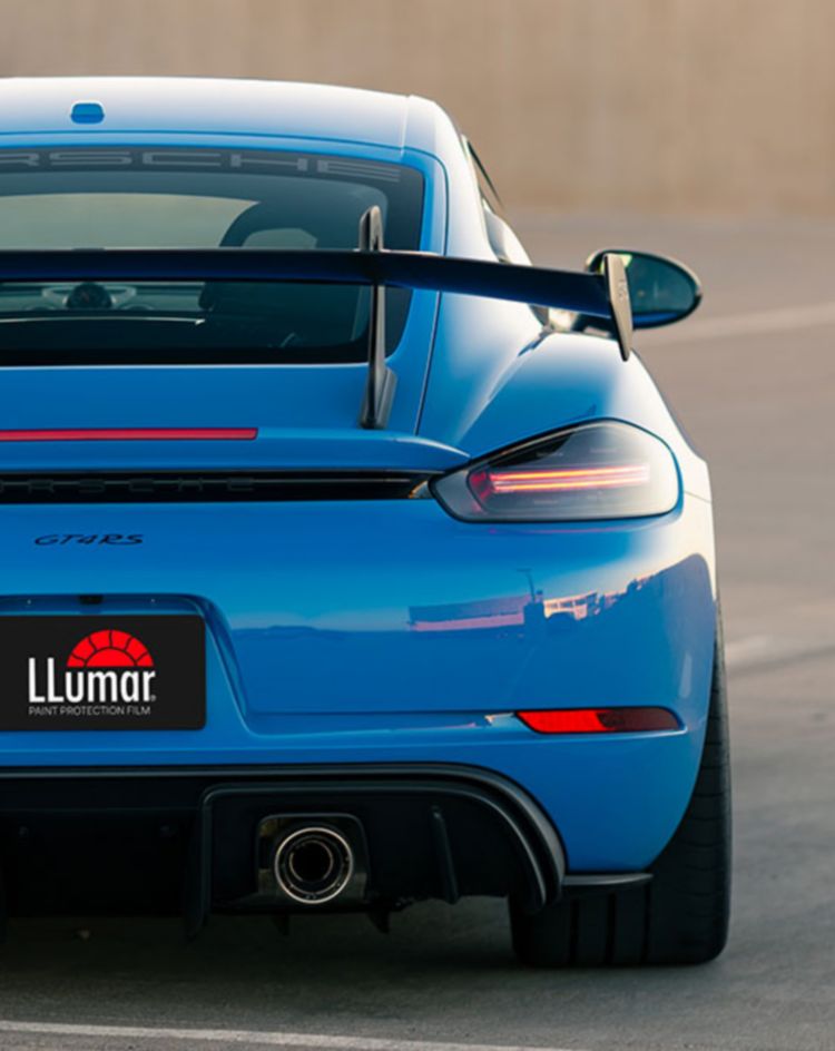 The rear view of a blue sports car with a large spoiler and dual exhausts, parked on a rooftop lot. 