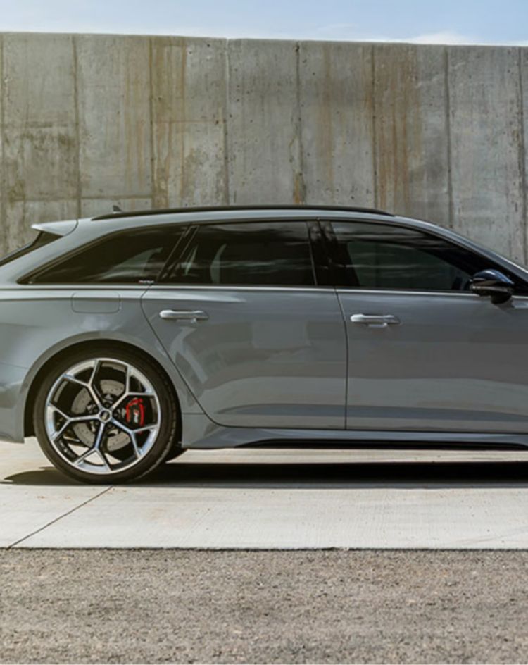 A gray performance wagon parked beside a tall concrete wall, shown from a side profile under bright daylight. 