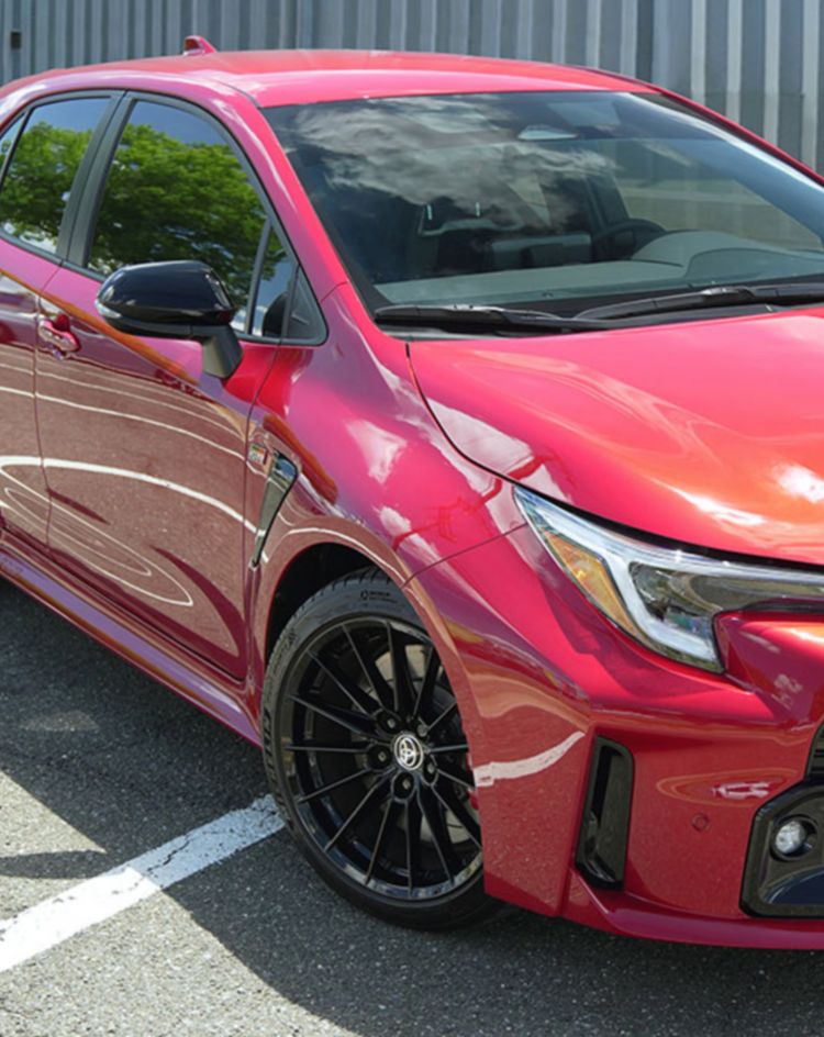 A red hatchback parked in an open lot with white parking lines and a metal-paneled building in the background. 