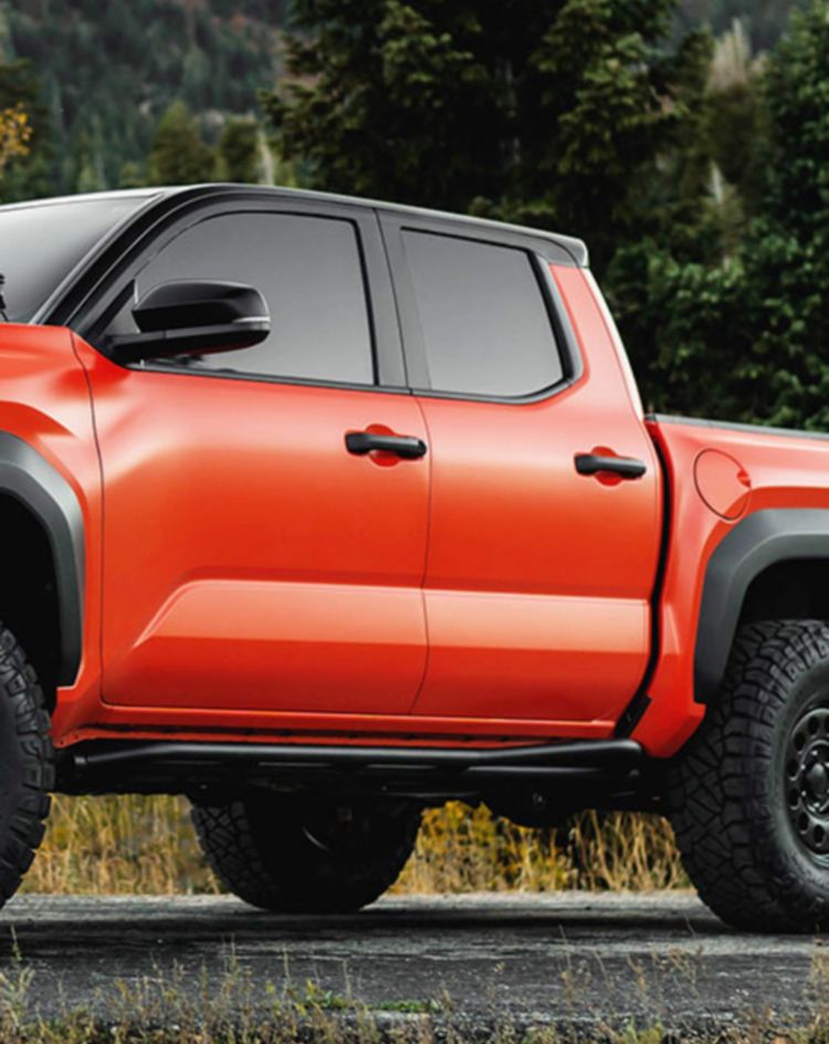 An orange off‑road pickup truck parked on a paved roadside with a backdrop of dense green and yellow autumn trees and mountain slopes. 