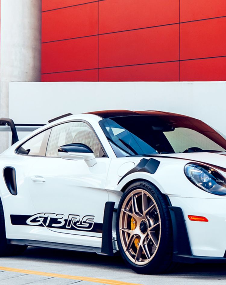 A white sports car with black accents and a large rear wing is parked on a modern concrete surface in front of a red and white building facade. 