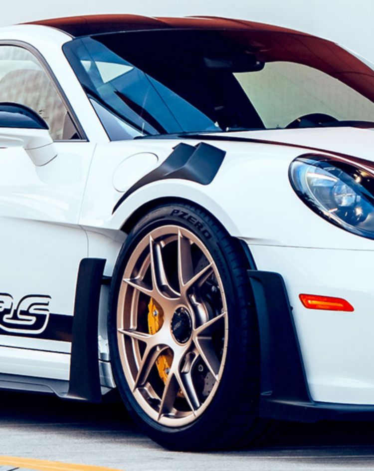 A white sports car with black accents and a large rear wing is parked on a modern concrete surface in front of a red and white building facade. 