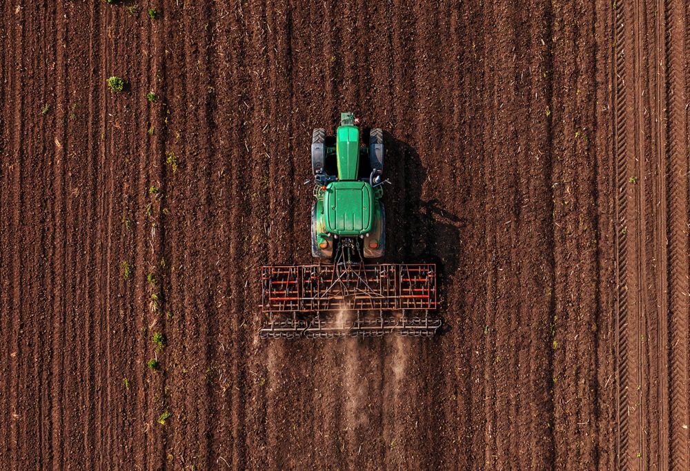 An aerial view of a green tractor tilling soil in a large field. 