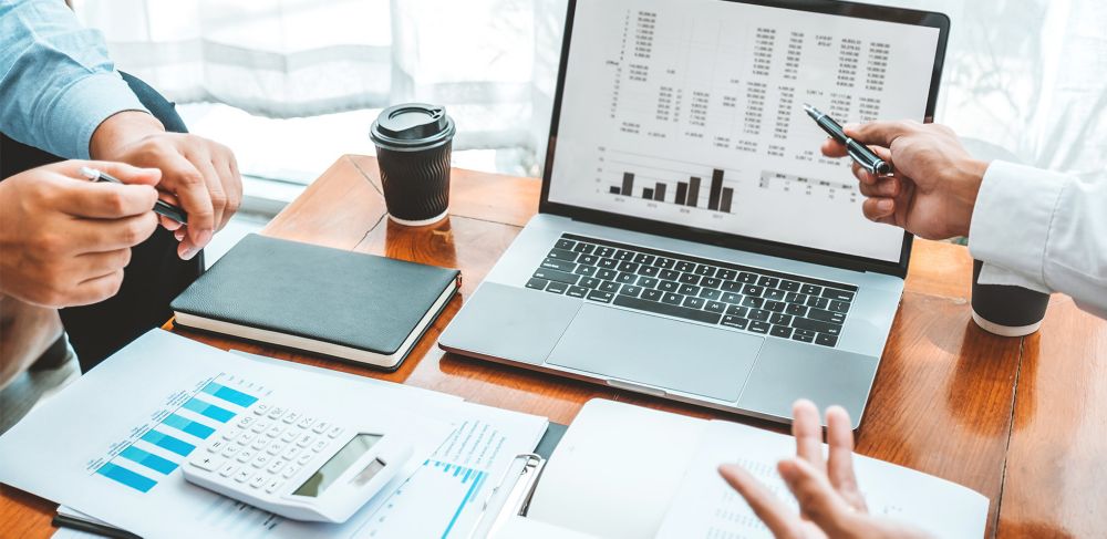 Two corporate executives reviewing financial charts on a laptop, surrounded by documents, calculator, notebook, and coffee on a wooden table. 