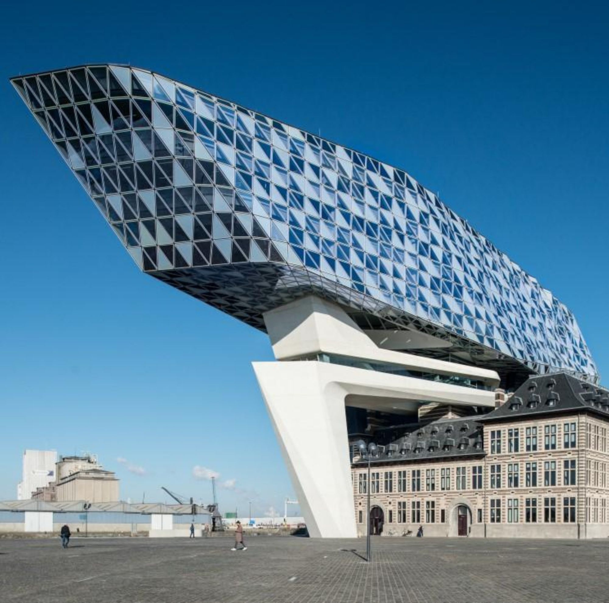 A modern,  glass building with a geometric facade rises above a historic square under a clear blue sky. 