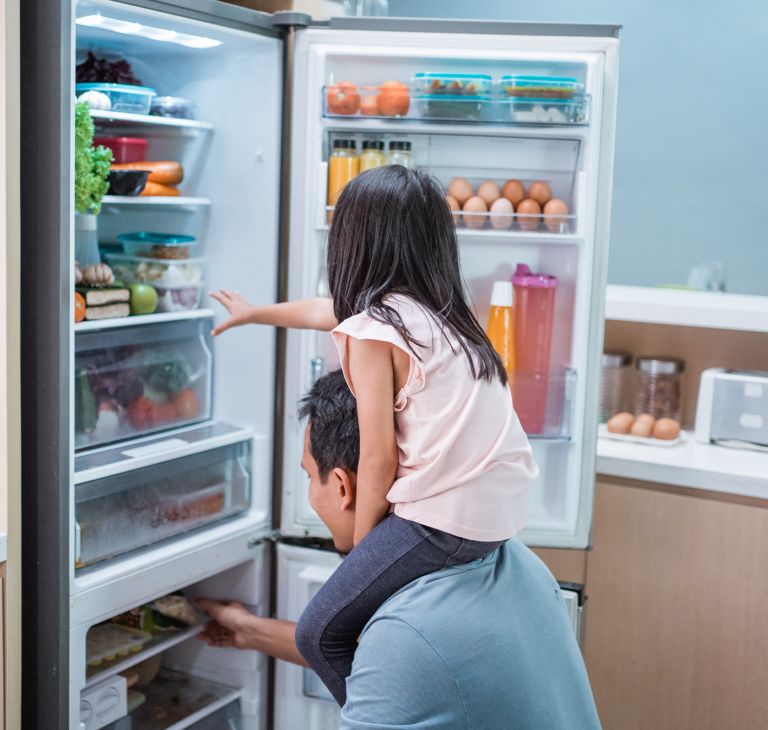 A child sits on their parent’s shoulders to look in the refrigerator. 