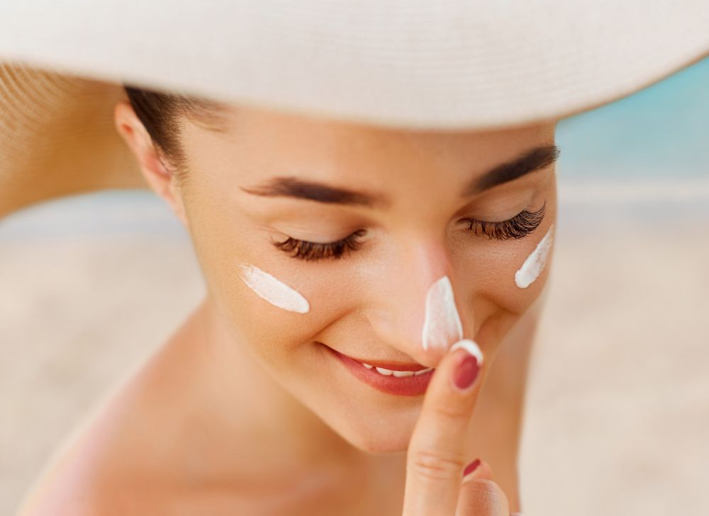 Woman on a beach applying sunscreen to her face while wearing a wide-brimmed floppy hat. 