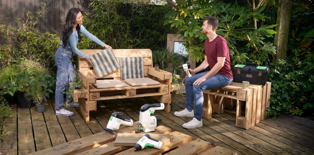 Two people enjoy outdoor patio furniture made of wood with various power tools and a toolbox nearby 
