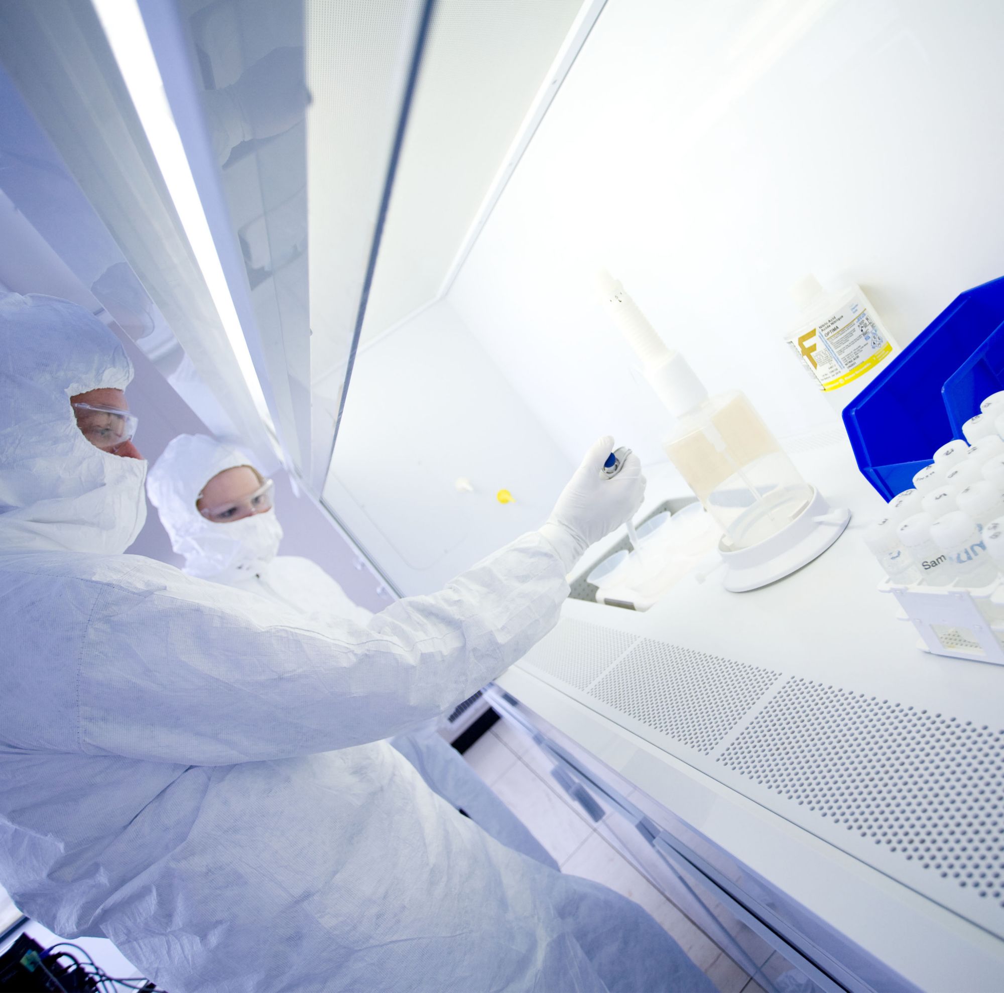 Scientists covered in personal protective equipment drop liquid into a container 