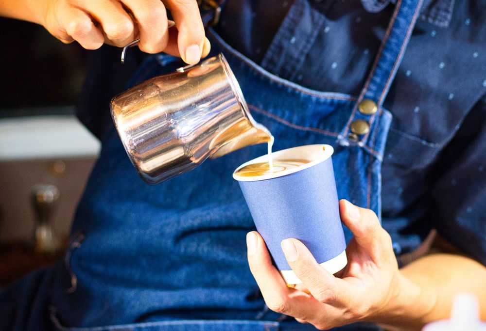 A barista wearing a blue apron pours creamer into a blue paper cup. 
