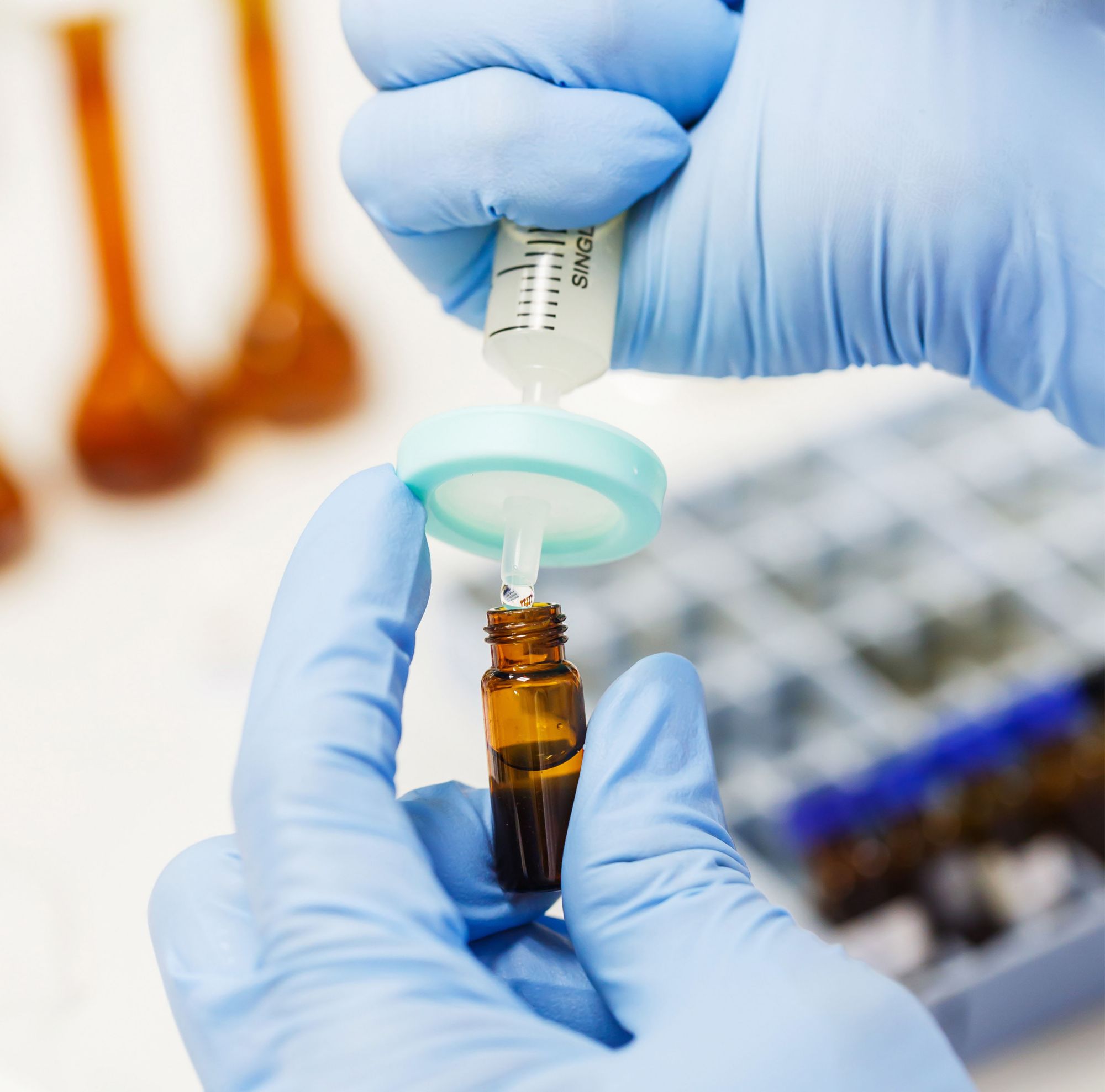 Someone wearing blue gloves injects a liquid into a brown vial in a lab. 