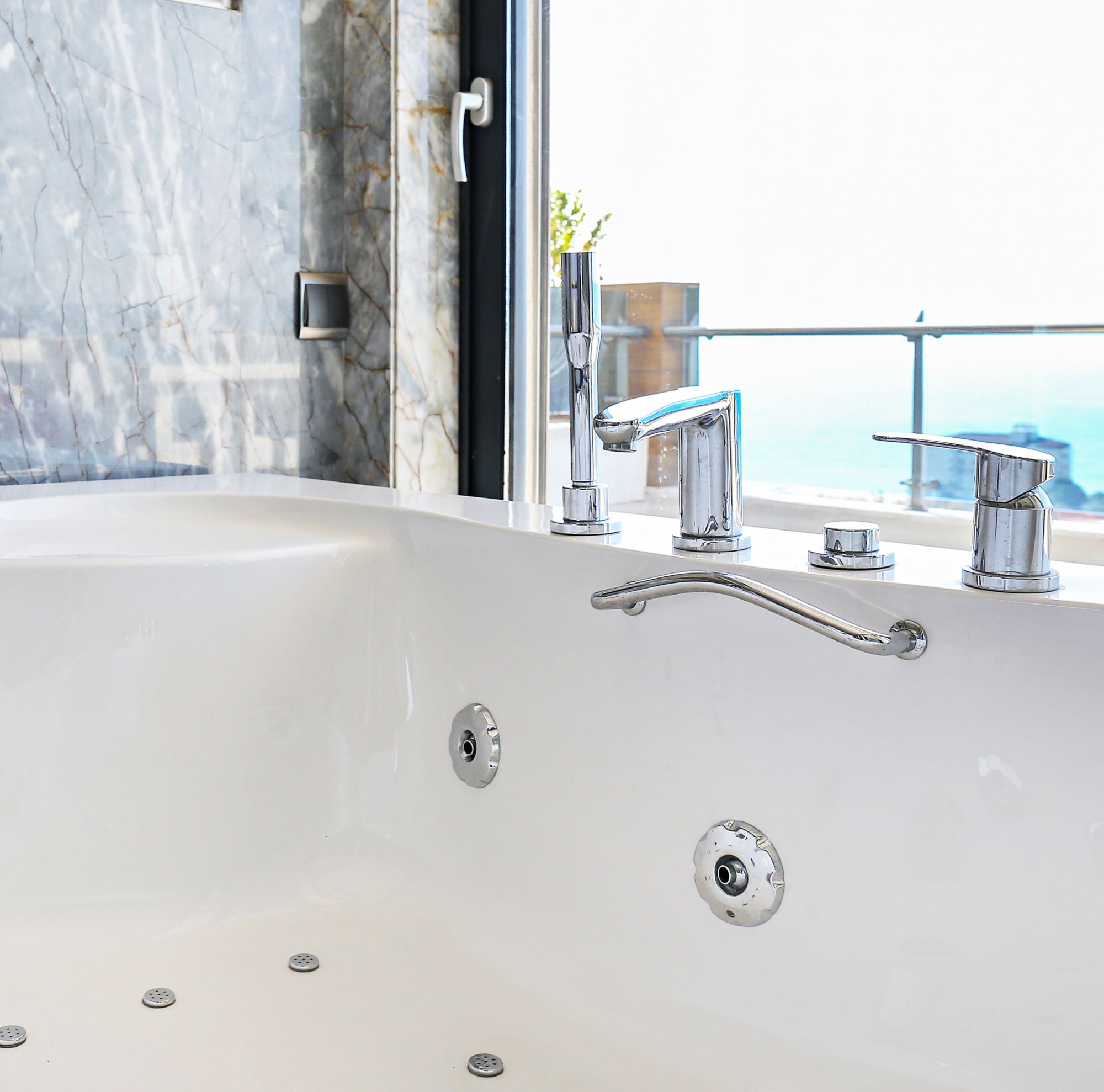 An empty white hot tub rests next to a window. Several small green plants are set around the tub.. 