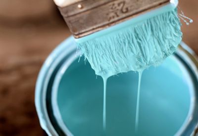 Light blue paint drips off a paintbrush into a paint bucket. 