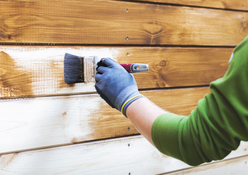 A gloved person brushes wood stain onto an exterior wood surface. 
