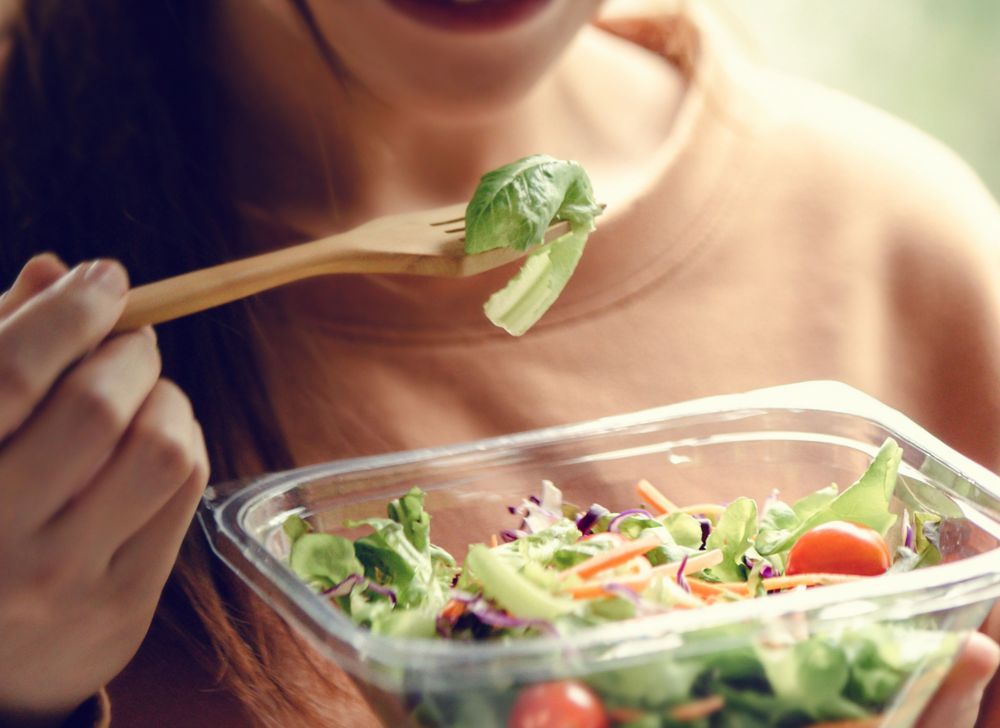 A person raises a forkful of salad from a container toward their mouth 