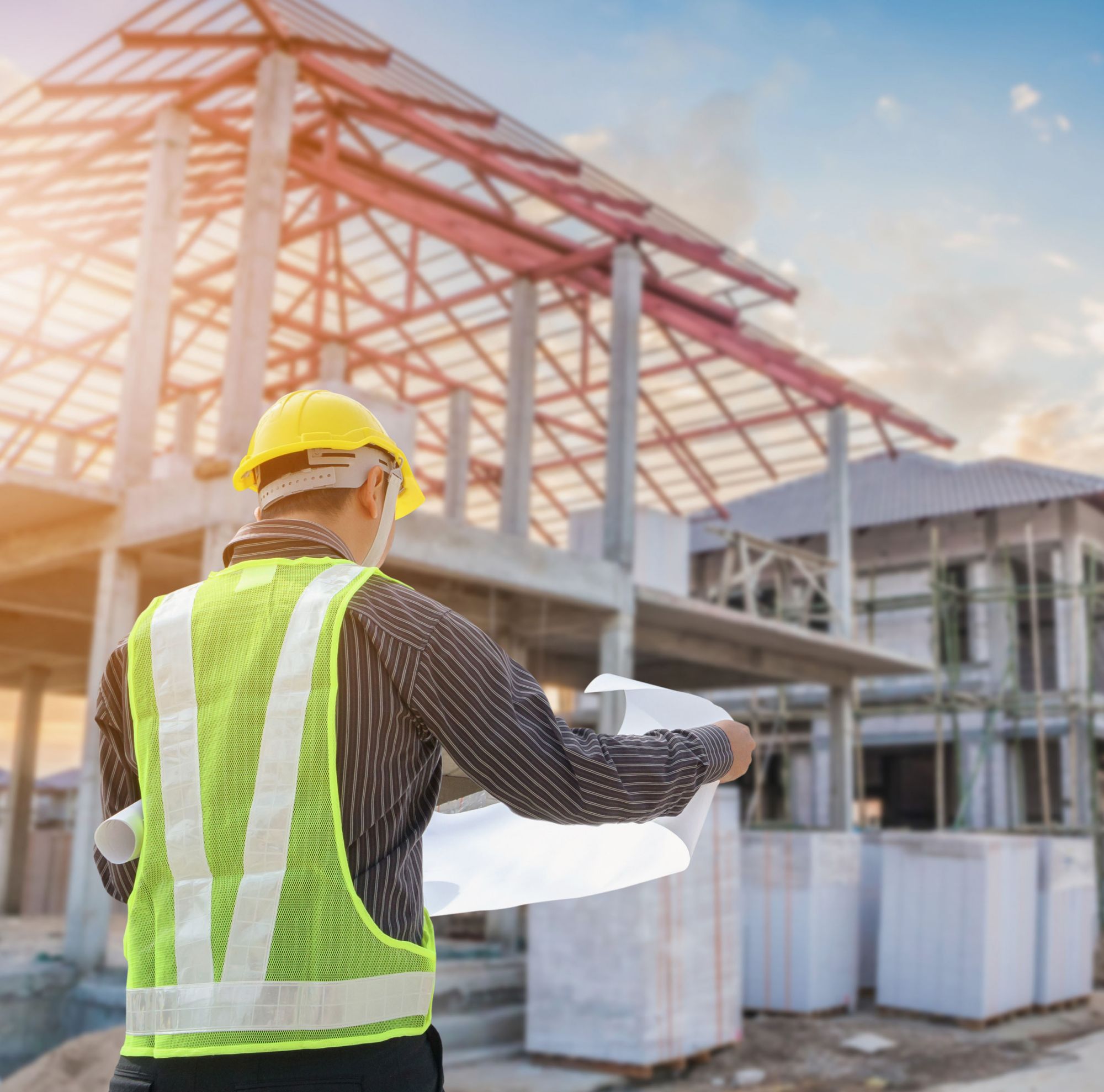 A person in a hard hat and vest holds blueprints while looking at a construction site. 