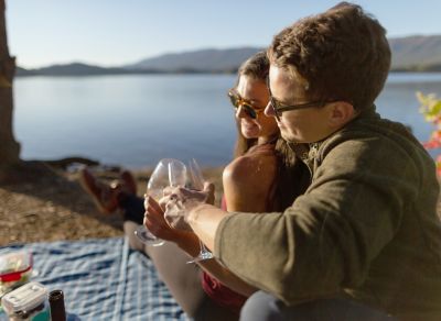 A man and woman cheers with wine glasses on a lake shore.