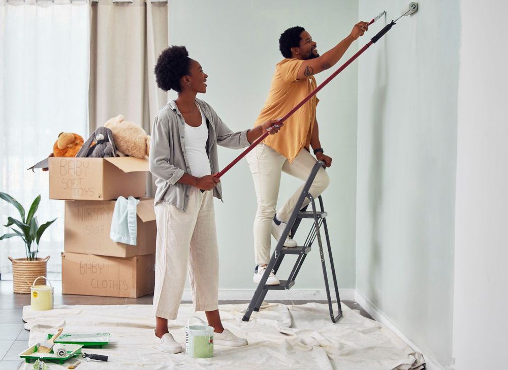 Two people are painting a wall lime green. One is standing on a tarp, and the other is standing on a step ladder that’s on the tarp. 