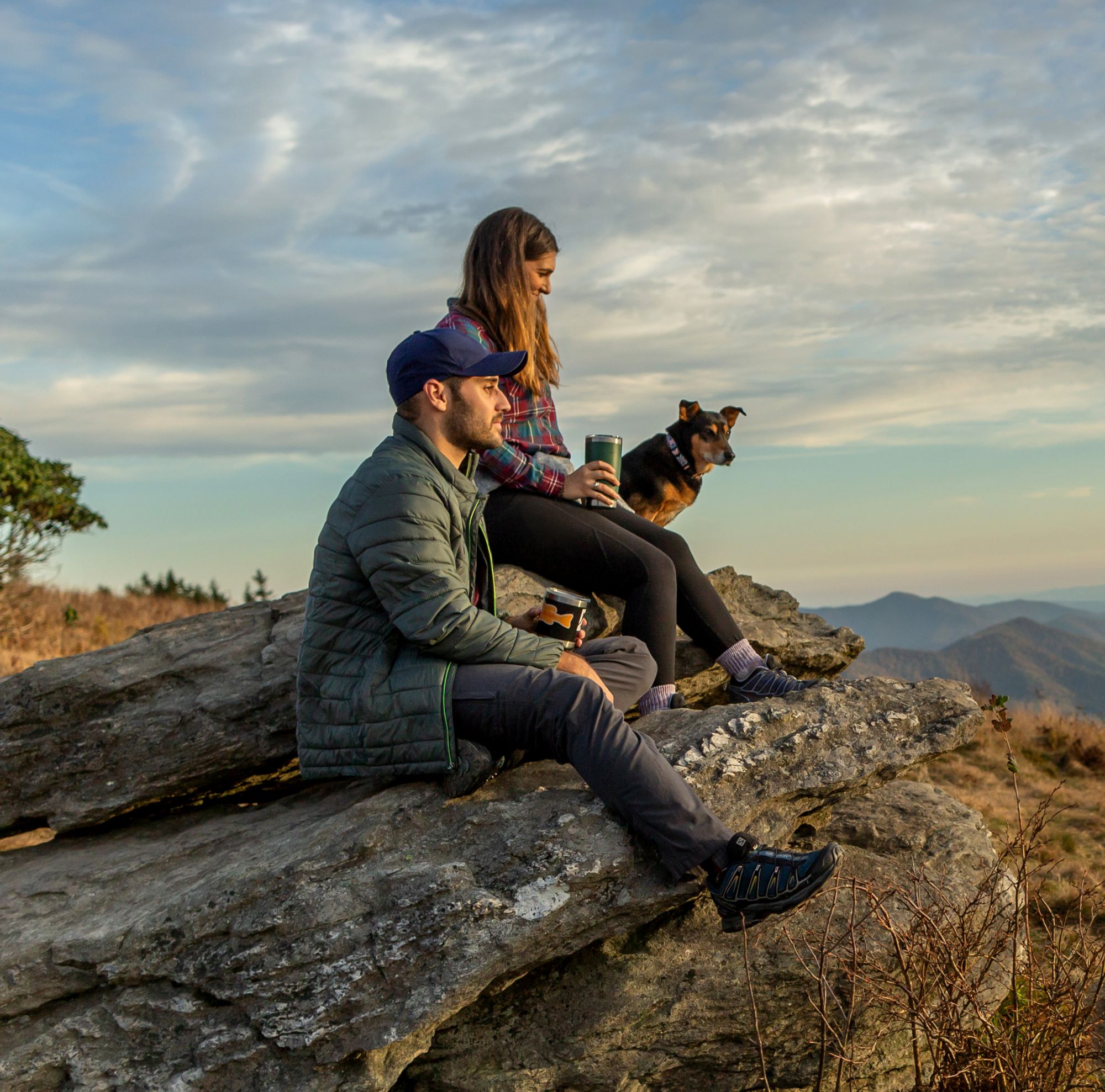 A man and woman hiking with their dog hold coffee cups and rest on a rock. 