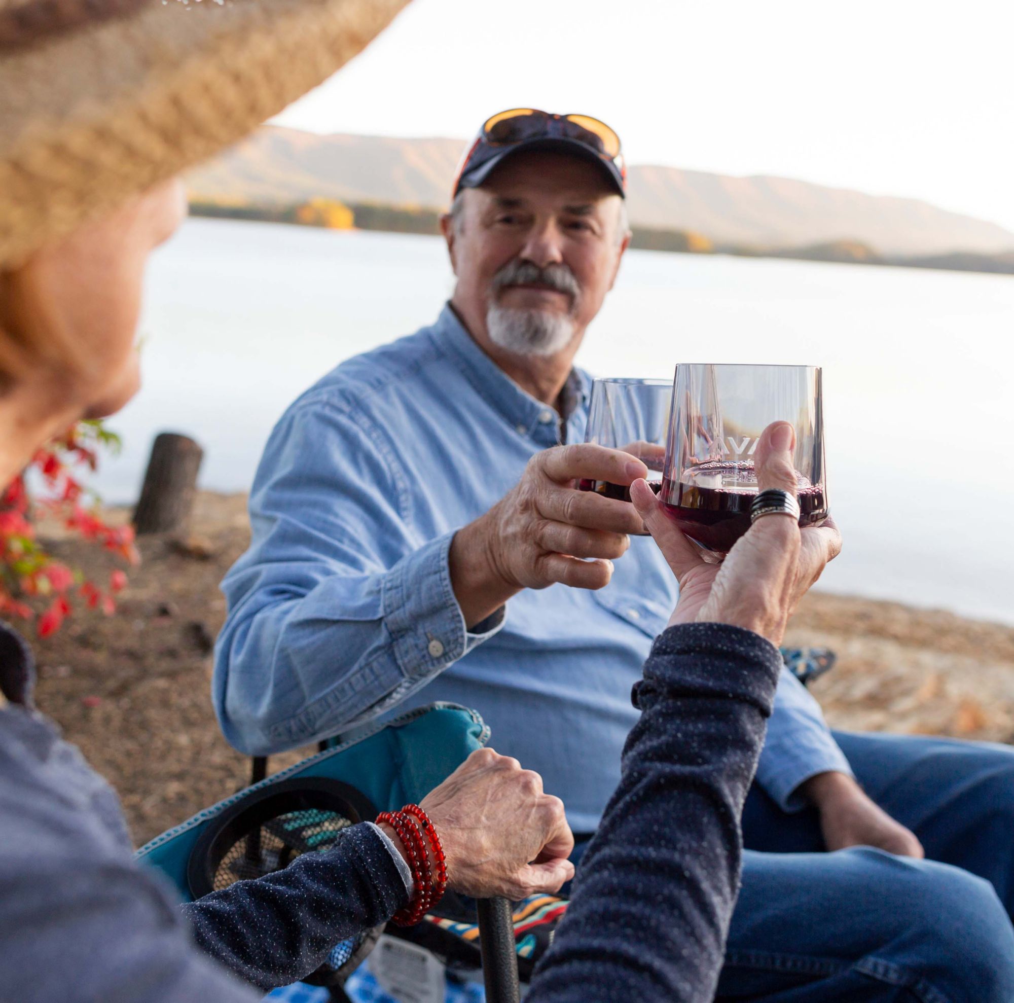 A couple sitting by a lake clinking their wine-filled, glass-like cups together. 