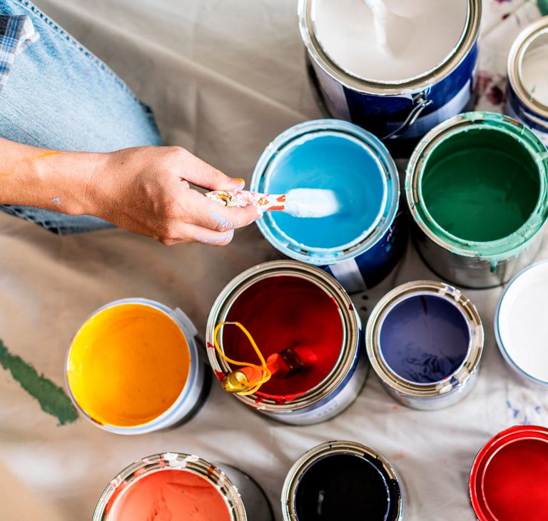 Hand dipping a brush in a can with blue paint among other cans of paintings. 