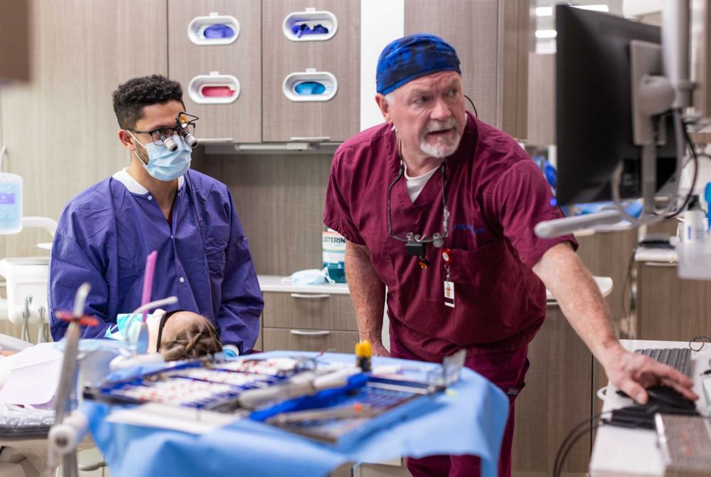 Dr. John Royal shows a student at the clinic notes on a computer screen while a patient sits in the dental chair.  