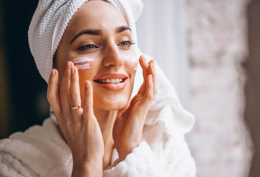A person with their hair in a towel applies white moisturizer to their cheeks.   