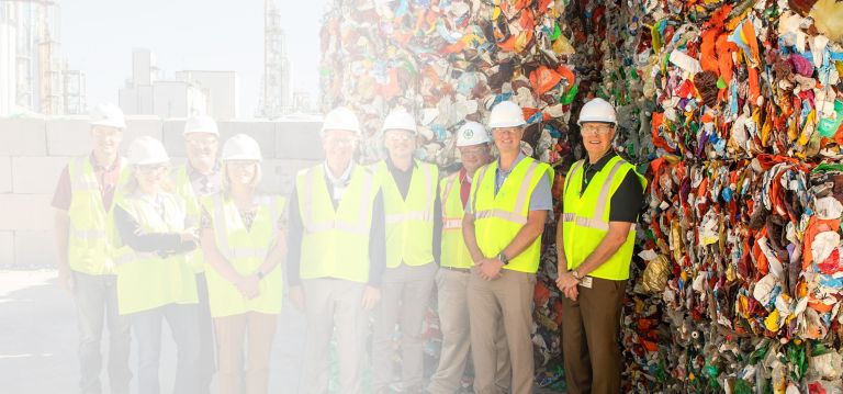 A group of people in safety vests and hard hats stand near baled plastic. 
