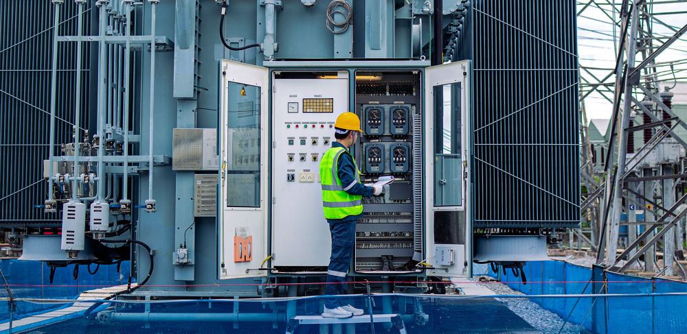 Engineer performs inspection on large transformer at electrical substation wearing safety gear and yellow helmet. 