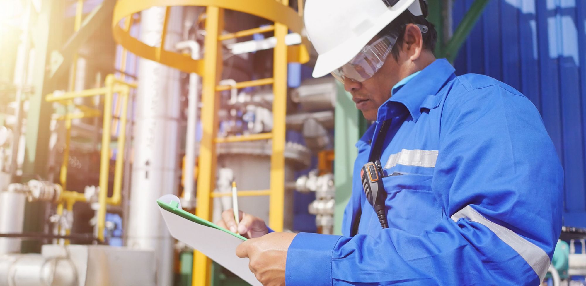 An engineer wearing proper PPE outside an industrial plant with a checklist in hand. 