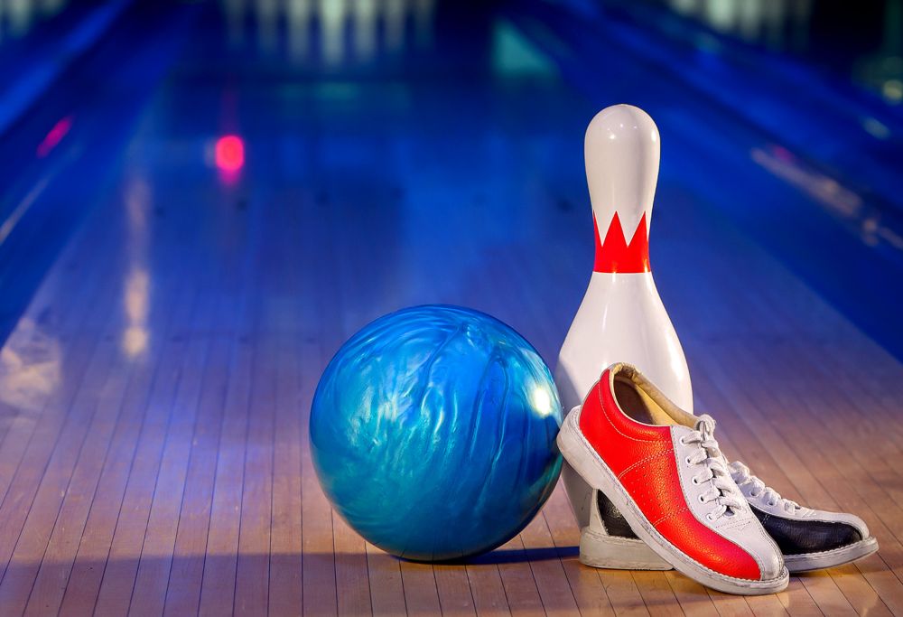 A bowling ball, pin and pair of shoes sit in front of a bowling lane. 