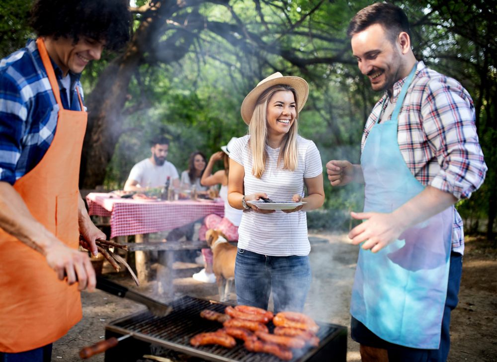 A man grilling hot dogs as others laugh around the grill. 