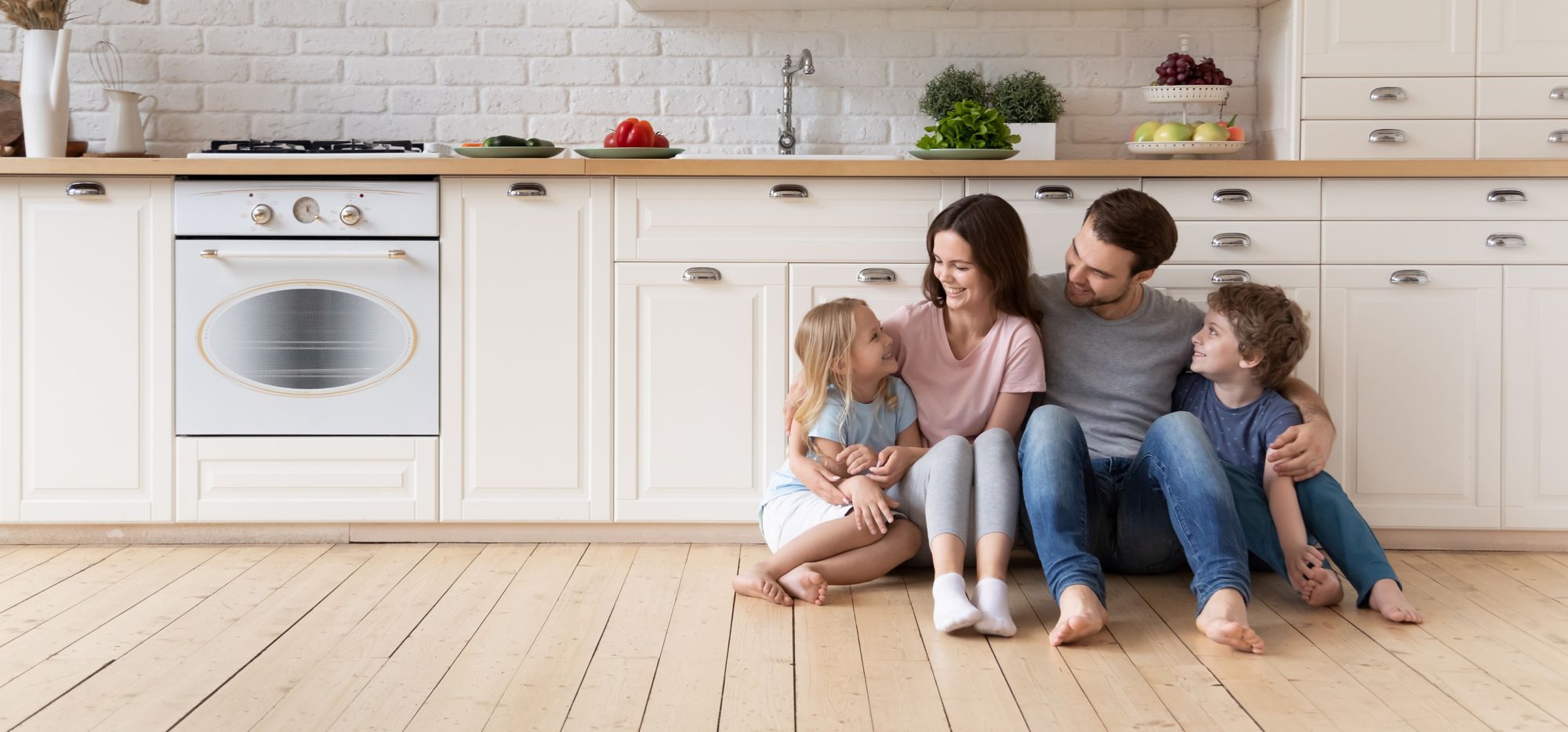 A family of four sits on their kitchen floor smiling and hugging each other. 