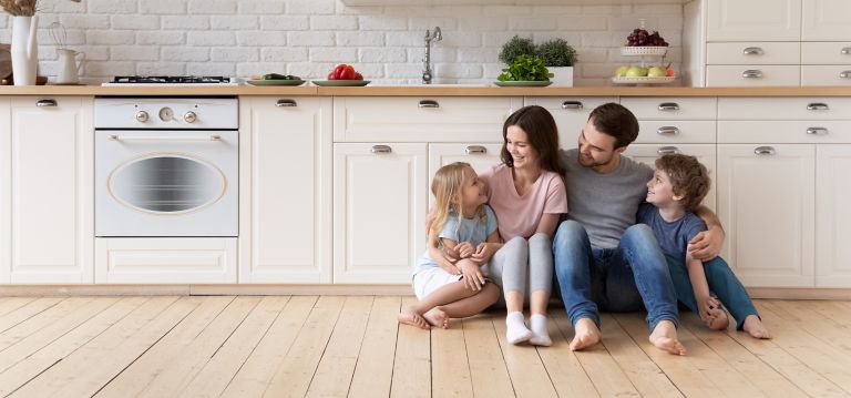 A family of four sit on their kitchen floor smiling. 
