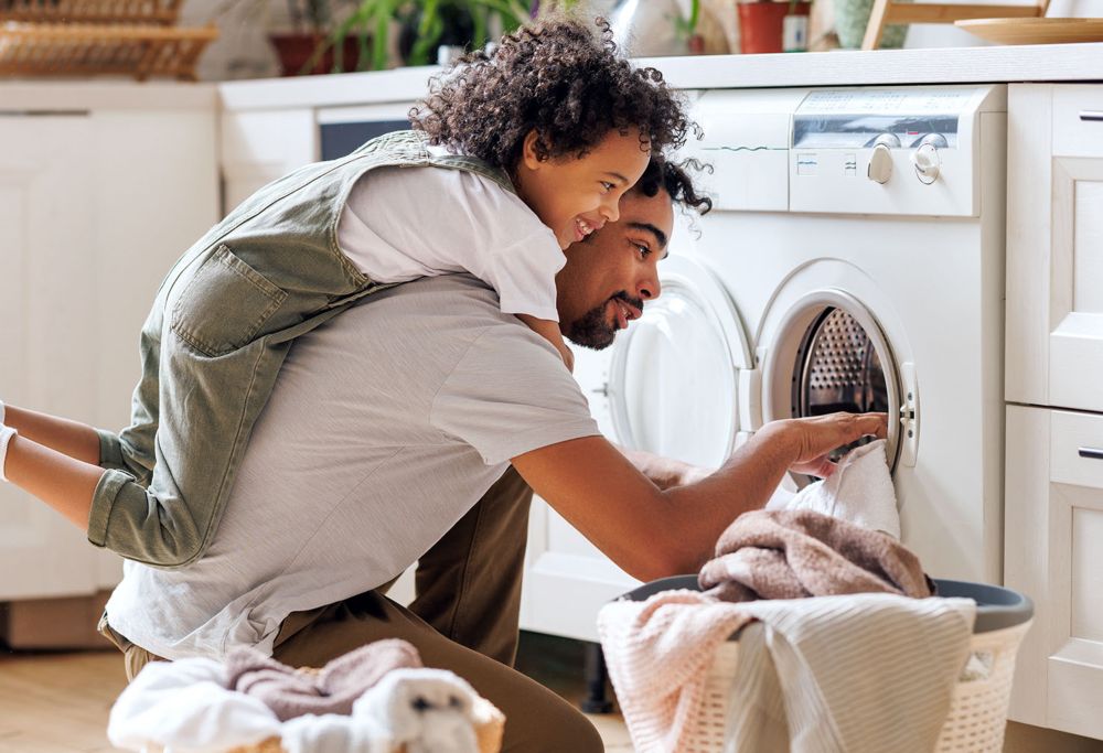 A father giving his son a piggyback ride as he puts clothes into the washer. 