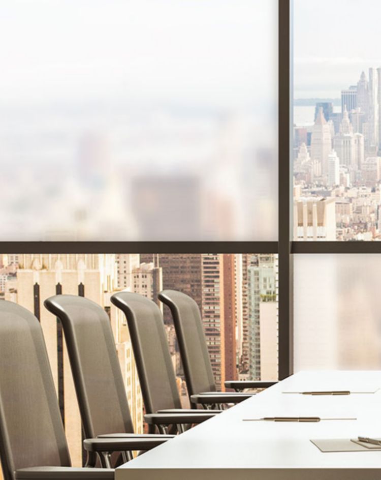 Large conference room featuring a long table and high‑back chairs, positioned in front of floor‑to‑ceiling windows showing a city skyline with tall buildings. 