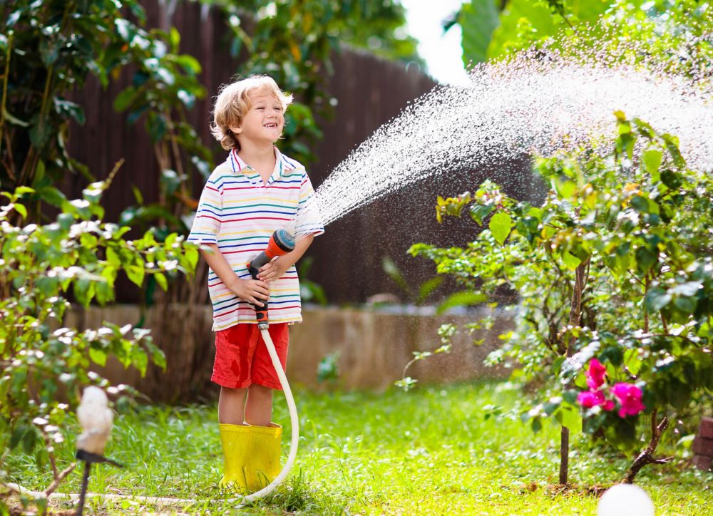 A boy waters plants while wearing rain boots in a yard. 